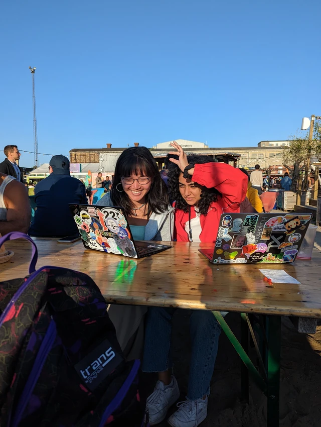 Two students lean together over sticker-covered laptops at an outdoor table in Copenhagen’s Reffen street-food market.