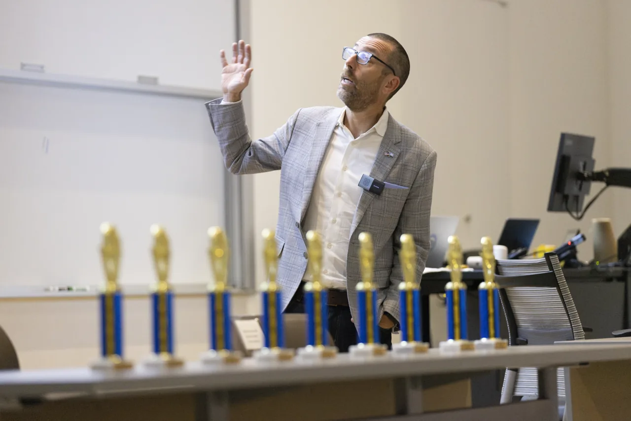 A speaker in a light gray suit giving the robotics seminar, with race-night trophies in the foreground